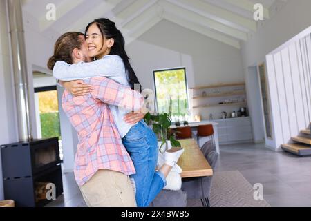 Coppie diverse che abbracciano la cucina casalinga luminosa. Lei ha lunghi capelli neri e lui sfoggia capelli corti marroni, entrambi vestiti casualmente, inalterati Foto Stock
