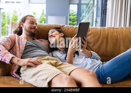 Coppie diverse che si rilassano sul divano di casa, guardando un tablet. Ha i capelli scuri, ha i capelli marrone chiaro e la barba, entrambi vestiti casual, inalterati Foto Stock