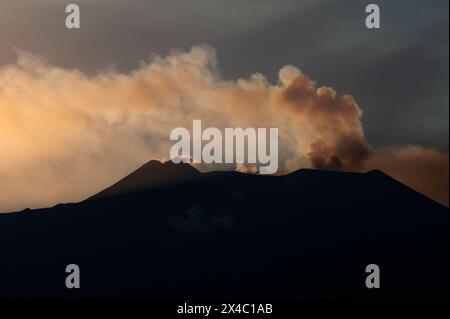Vista distante dall'est dell'Etna in Sicilia, Italia. I pennacchi di fumo e le nuvole di gas solforoso provenienti dai crateri sommitali sono retroilluminati dai raggi del sole che tramonta. Foto Stock
