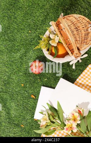 Un cestino di bambù con frutta e fiori decorato con quaderni bianchi e tessuto a scacchi su sfondo verde. Vista dall'alto, spazio di copia per Foto Stock