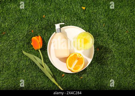 Vista dall'alto di un contenitore rotondo per piatti bianchi, una bottiglia cosmetica con tiralatte, mezza tazza di arancia e frullato decorata con fiori di tulipani su un backgrou di erba verde Foto Stock