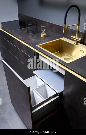 Armadio del cestino della cucina. Estrarre il cassetto della cucina per il cestino raccolta differenziata sotto il lavabo dorato e il rubinetto piano cottura elettrico in vetro nero Foto Stock