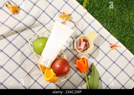 Cestino da picnic in vimini con frutta, marmellata e fiori sull'erba verde del parco. Un modello vuoto in plastica bianca per cosmetici con spazio per il design pa Foto Stock