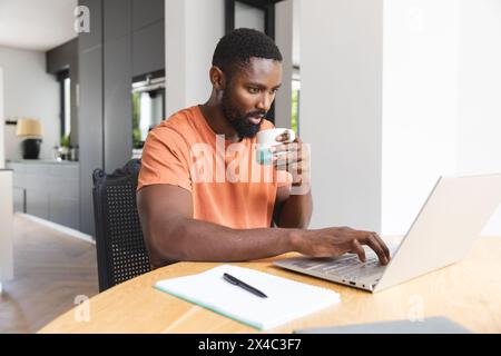 Proprietario di casa afroamericano maschio che usa il portatile a casa, tenendo la tazza. Ha la barba, indossa una camicia arancione, sembra concentrato, inalterato Foto Stock