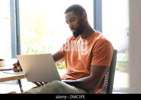 Proprietario di casa afroamericano seduto, che usa un computer portatile. Ha la barba, indossa una camicia arancione, concentrato sullo schermo, inalterato Foto Stock