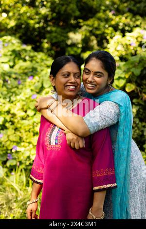 Madre indiana e figlia adolescente, entrambe con capelli scuri, abbracciate all'aperto. Indossa un kurta rosa, madre che tiene la figlia in blu, entrambi sorridenti, enjo Foto Stock