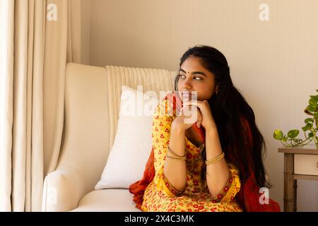 Giovane donna indiana che indossa un abbigliamento tradizionale, seduta con cura a casa, spazio fotocopie. Lunghi capelli neri e bracciale rigido in oro, circondati da lussureggianti Foto Stock