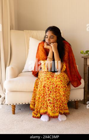 Donna indiana in abito giallo, con libro sul divano color crema, sciarpa arancione. Lunghi capelli neri che incorniciano il suo volto premuroso, la figlia adolescente accanto a h Foto Stock