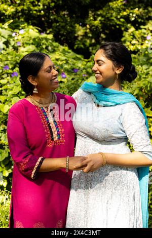 Madre indiana e figlia adolescente, che indossano abiti tradizionali e sorridono all'aperto. Condividere momenti gioiosi, espressioni che irradiano felicità e amore aga Foto Stock