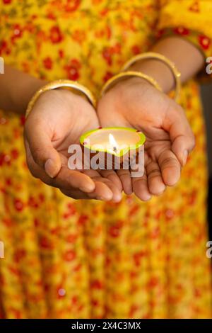 Una donna indiana che tiene accesa la candela e indossa un abito floreale. Unghie e braccialetti con vernice sportiva, in piedi su uno sfondo sereno, sorridendo dolcemente Foto Stock
