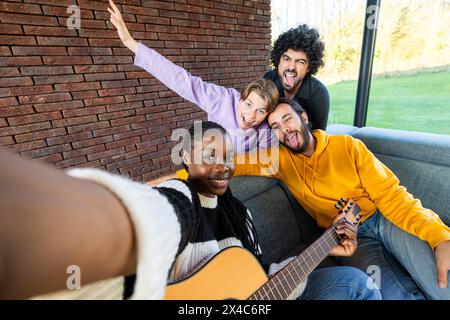 Cattura un momento di pura gioia, questa immagine mostra un gruppo di amici diversi che scattano un selfie. Uno suona una chitarra, mentre gli altri mostrano espressioni giocose. Sono sedute contro un muro di mattoni all'interno di una sala luminosa con grandi finestre che rivelano un lussureggiante sfondo verde all'aperto. Amici allegri e variegati che portano Selfie con Guitar indoor. Foto di alta qualità Foto Stock