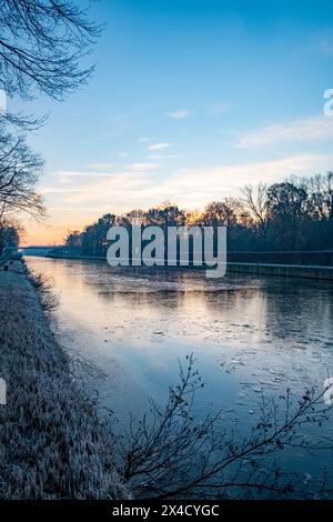 Questa immagine cattura la bellezza serena di un'alba invernale, con la luce precoce che proietta una luce soffusa su un fiume calmo. La superficie dell'acqua riflette il cielo risveglio, mentre frammenti di ghiaccio fluttuano silenziosamente, segnando il freddo abbraccio della stagione. Le sagome degli alberi senza foglie fiancheggiano la riva del fiume, in piedi contro la luce dell'alba, e le erbe smerigliate lungo il bordo aggiungono un dettaglio nitido al paesaggio fluviale. Questo momento di tranquillità è un rinnovamento quotidiano, una promessa della luce e del calore che verranno dopo la lunga notte. Dawn's Reflection: L'alba invernale sul fiume. Foto di alta qualità Foto Stock