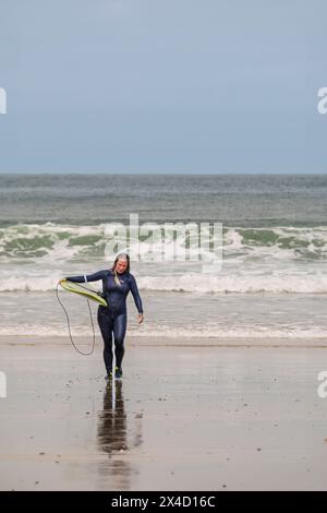 Una surfista matura che porta la sua tavola da surf e cammina fuori dal mare a Towan Beach a Newquay in Cornovaglia nel Regno Unito. Foto Stock