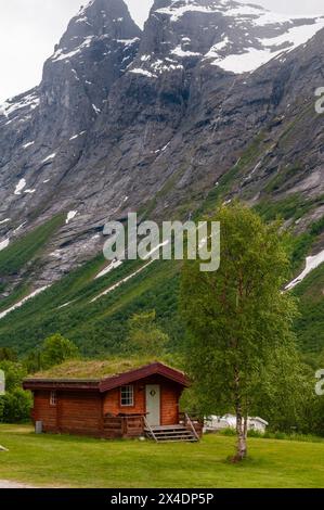 Una capanna di legno primitiva annidata in una valle verdeggiante alla base di scogliere a strapiombo ricoperte di ghiaccio. Trollstigen, Rauma, Norvegia. Foto Stock