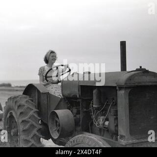 anni '1950, storica, fuori su una spiaggia, una giovane donna a metà anni '20, seduta su un vecchio trattore, che tiene il grande volante in metallo. Grazie al potente motore e alle grandi ruote posteriori, i trattori sono comunemente visti sulle spiagge dove vengono utilizzate alghe limpide, riordinare la sabbia e trainare le barche. Foto Stock