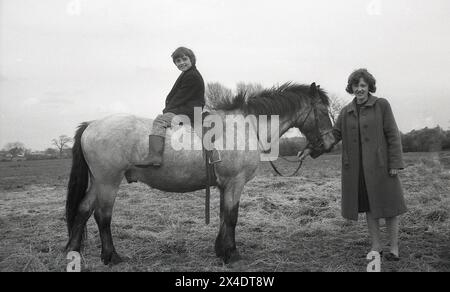 anni '1960, storica, all'esterno di una scuola di equitazione suburbana, una giovane ragazza seduta a cavallo, mani sulla testa, sua madre che tiene le redini del cavallo. Sedersi in questo modo su un cavallo aiuta a imparare a bilanciarsi durante la guida. Foto Stock