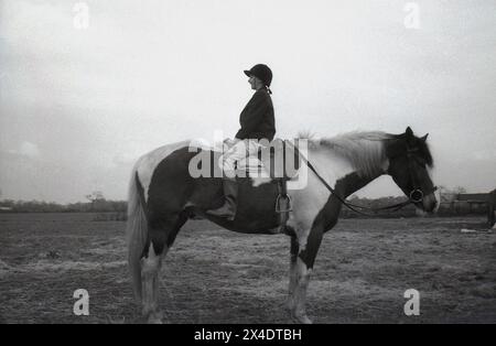 anni '1960, storica, fuori in un campo, in una scuola di equitazione suburbana, una ragazza seduta a cavallo, qualcosa fatto per migliorare il suo equilibrio durante la guida. Foto Stock