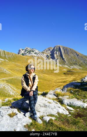 Una ragazza sorridente siede su una pietra in una pittoresca area montuosa e ammira la vista. Sullo sfondo del Monte Prutas, il Parco Nazionale Durmitor Foto Stock