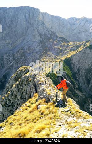 Un uomo con uno zaino cammina lungo un pericoloso crinale, aggrappandosi a una corda di sicurezza, una vacanza attiva tra le pittoresche montagne Foto Stock