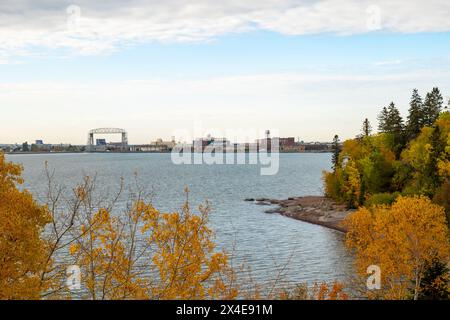 Vista dell'iconico ponte aereo Duluth Minnesota Aerial Lift Bridge, dell'acqua e della riva del lago Superior, delle vicine foglie autunnali e degli edifici del porto distanti su un'area di lavoro nuvolosa Foto Stock