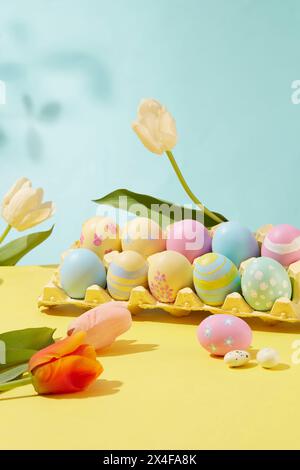 Gioia pasquale catturata in una cornice pittoresca. Le uova colorate e fantasia sono disposte in un blister decorato con tulipani su sfondo color pastello. Foto Stock