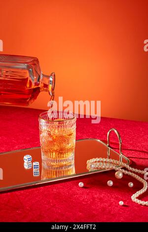 Il whisky viene versato in un bicchiere da un decanter. Vista frontale, tazza di whisky esposta sullo specchio con dadini e collana di perle su dorso arancione e rosso Foto Stock