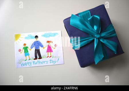 Festa del papà foto di una cartolina disegnata da un bambino. Un regalo di colore blu con un fiocco. Sfondo bianco. La cartolina mostra un padre che cammina con sua figlia e suo figlio. Buona festa del papà. Foto Stock