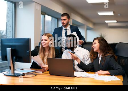 Il team multietnico lavora in un ufficio moderno, una giovane donna punta sullo schermo del computer, presentando idee. I colleghi si riuniscono intorno alla scrivania con un laptop e con i documenti Foto Stock