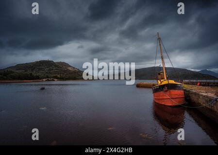 Vecchia barca legata su un molo troppo coltivato a Invergaray / Loch fine. Sullo sfondo l'Aray Bridge. Foto Stock