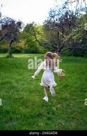 una bambina dai capelli lunghi corre nel giardino con un cestino pieno di fiori Foto Stock