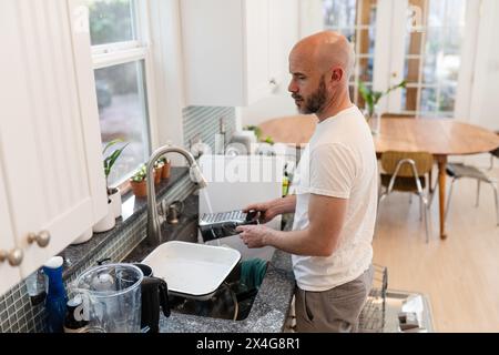 Uomo che sciacqua i piatti in una cucina soleggiata Foto Stock