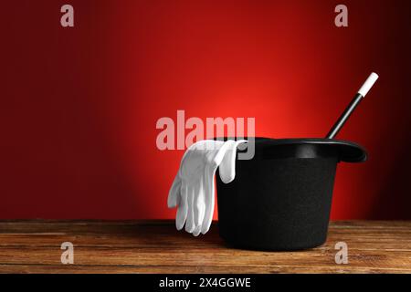 Cappello, guanti e bacchetta da mago su un tavolo di legno su sfondo rosso, spazio per il testo Foto Stock