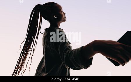 Una giovane donna sta in uno studio, tenendo un telefono cellulare a occhi chiusi e braccia tese. Ascolta la musica tramite auricolari wireless. Foto Stock