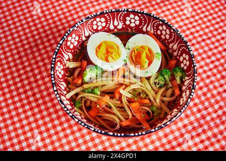 Sana zuppa di noodle di ramen in stile asiatico con verdure e uova perfettamente cotte, servita in una ciotola decorativa su tovaglia a scacchi rossi Foto Stock