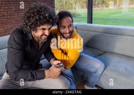 In una camera piena di luce con vista sul verde all'aperto, due amici condividono un divano e un momento di pura gioia. L'uomo con la camicia nera regge uno smartphone, forse condividendo qualcosa di umoristico mentre entrambi si appoggiano, ridendo. L'altro, in una felpa gialla luminosa, esprime la gioia, contribuendo all'atmosfera vivace e spensierata. Risate condivise: Due amici che si godono un momento di cuore leggero sul divano. Foto di alta qualità Foto Stock