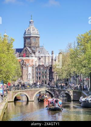 Amsterdam Paesi Bassi 21 aprile 2024 Una barca con turisti scivola tranquillamente lungo un fiume lungo il canale accanto a un imponente e maestoso edificio, creando un contrasto sorprendente tra paesaggi naturali e urbani. Foto Stock