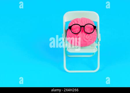 Modello di cervello umano, adornato da occhiali, seduto su una sedia pieghevole di tè, isolato su uno sfondo blu con spazio di copia. Competenza medica e medico r Foto Stock