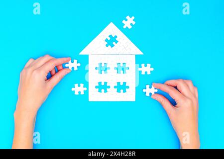 Mani che assemblano un puzzle a forma di casa con un tetto all'aperto, su uno sfondo blu. Foto Stock