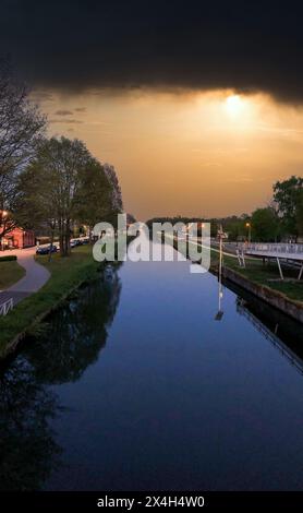 Mentre il crepuscolo scende, un canale si estende in lontananza, fiancheggiato da alberi e dal caldo bagliore delle luci della strada. Il sole tramonta attraverso le pesanti nuvole, gettando una luce spettacolare sull'acqua tranquilla. Questa pittoresca scena cattura la tranquilla transizione dal giorno alla notte in una tranquilla città sul mare. Majestic Canal al crepuscolo con luci di strada scintillanti. Foto di alta qualità Foto Stock