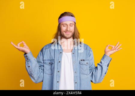 Ritratto fotografico di un giovane uomo hippie che medita gli occhi chiusi indossa abiti alla moda in denim isolati su sfondo giallo Foto Stock