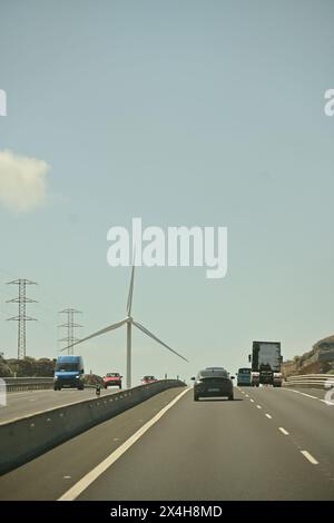 autostrada trafficata con auto, turbina eolica e linee elettriche, che catturano il contrasto tra tecnologia moderna e infrastruttura tradizionale Foto Stock