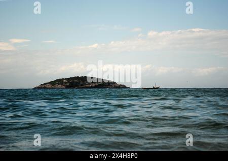 isola in lontananza, circondata da calme acque oceaniche e da un cielo azzurro, con una piccola barca che si aggiunge alla tranquilla scena Foto Stock