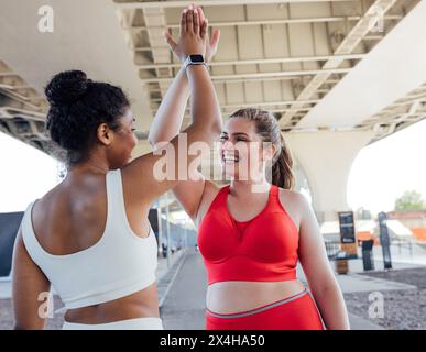 Due femmine più grandi si danno cinque all'aperto. Donna felice in abbigliamento sportivo rosso che esulta con la sua amica durante un allenamento. Foto Stock