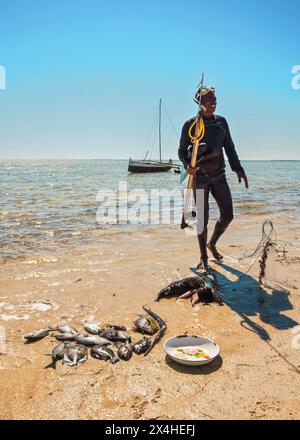 Anakao, Madagascar - 3 maggio 2019: Pescatore malgascio locale in muta in neoprene, con snorkeling e arpione semplice, in piedi accanto al mucchio di caugh fresco Foto Stock