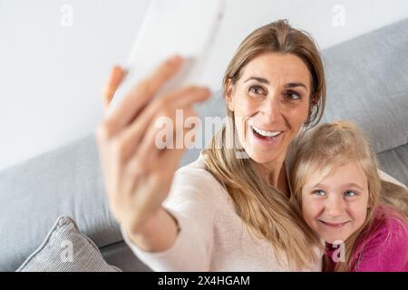 Madre e figlia felici sorridono mentre si fanno un selfie su un divano grigio, luminosi e allegri Foto Stock