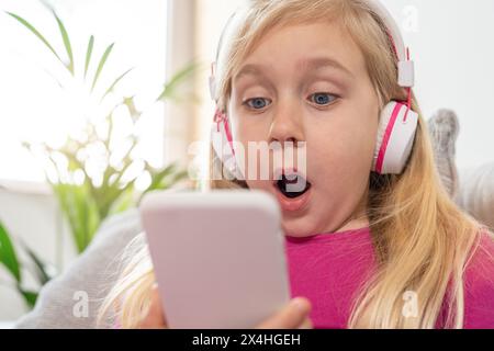 La bambina con una camicia rosa e le cuffie è scioccata e sorpresa da ciò che vede mentre fissa il suo telefono cellulare Foto Stock