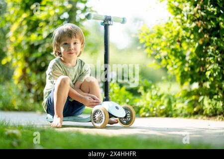 Adorabile bambino in sella al suo scooter in un cortile posteriore nella soleggiata sera d'estate. Bambino piccolo in sella a un rullo. Attività ricreative e sport all'aperto per i bambini Foto Stock