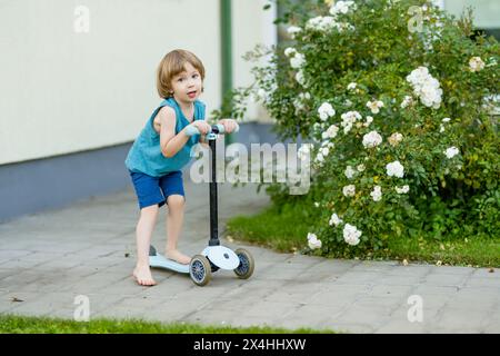 Adorabile bambino in sella al suo scooter in un cortile posteriore nella soleggiata sera d'estate. Bambino piccolo in sella a un rullo. Attività ricreative e sport all'aperto per i bambini Foto Stock