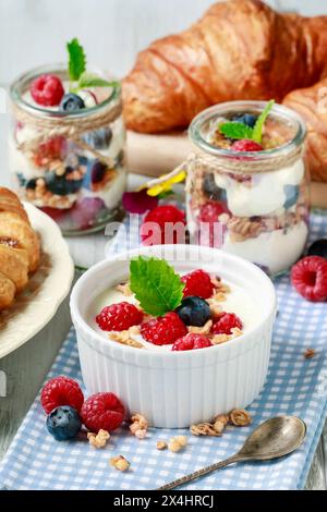 Tavolo della colazione: Ciotola di yogurt con muesli e frutta fresca: Lamponi e mirtilli, vasetti di vetro con frutta e panna montata. Foto Stock