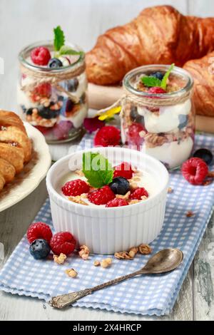 Tavolo della colazione: Ciotola di yogurt con muesli e frutta fresca: Lamponi e mirtilli, vasetti di vetro con frutta e panna montata. Foto Stock